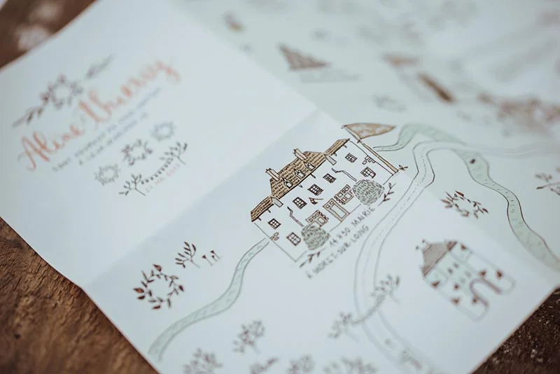 Gros plan sur un faire-part de mariage blanc avec une illustration aquarelle d'une maison et une calligraphie élégante.