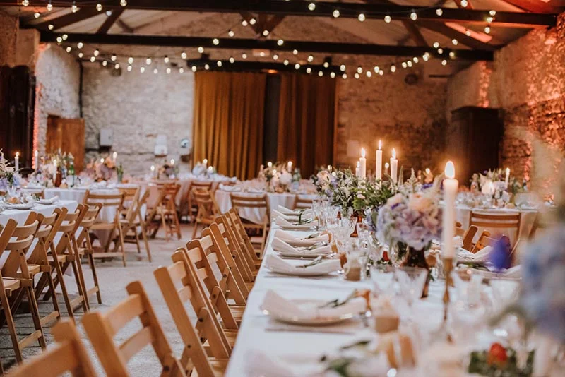 Réception de mariage chaleureuse dans une grange en pierre avec tables dressées, bougies et guirlandes lumineuses.