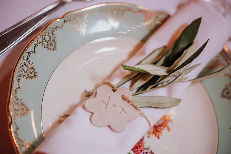 Détail d'une assiette vintage avec une serviette rose, un brin d'olivier et un marque-place en forme de fleur.