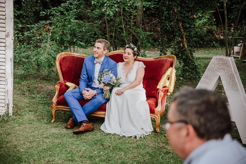 Jeunes mariés souriants assis sur un canapé rouge vintage en extérieur, entourés de verdure.