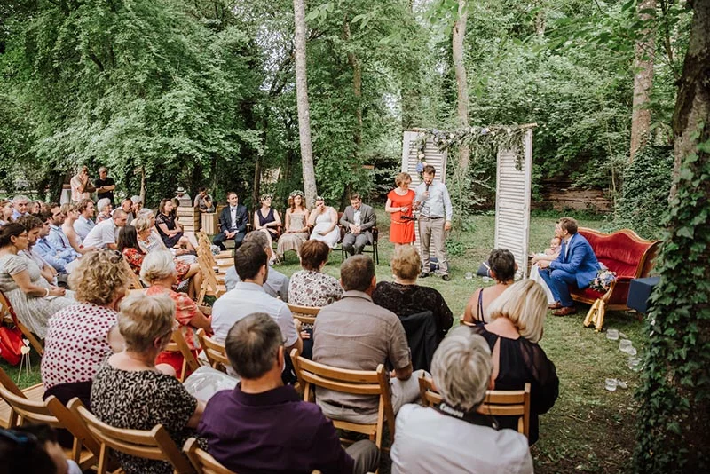Cérémonie de mariage champêtre en plein air avec invités assis face aux mariés sous une arche de volets blancs.
