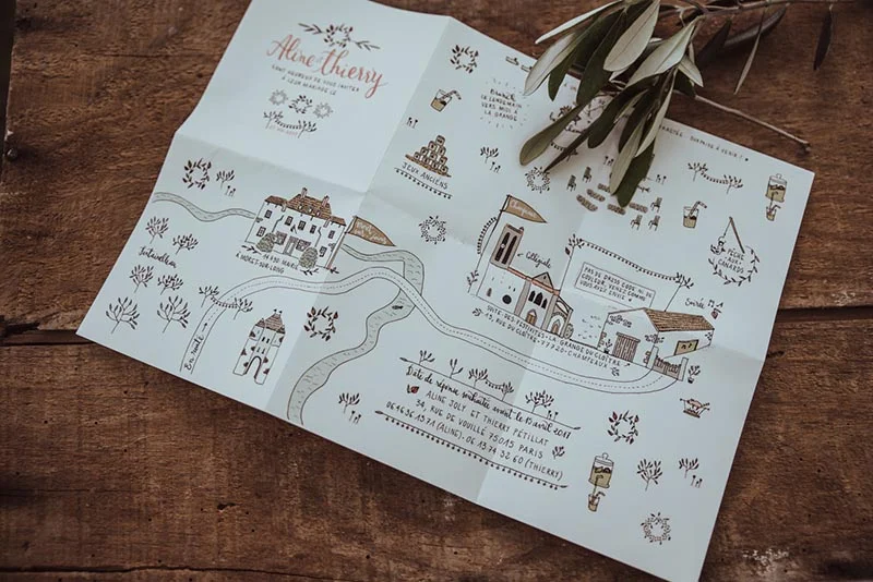 Faire-part de mariage illustré d'un plan dessiné à la main sur papier blanc, posé sur un fond en bois rustique.