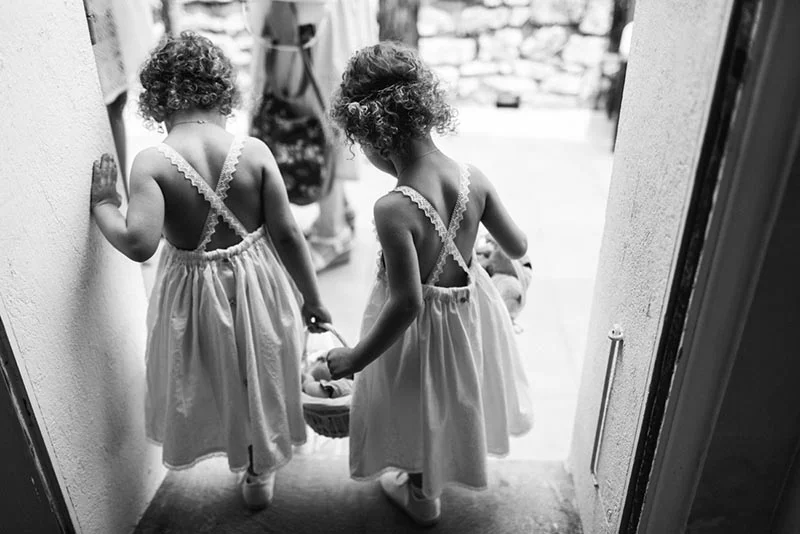 Deux petites filles en robes blanches et boucles blondes, vues de dos, s'apprêtent à entrer dans une pièce.