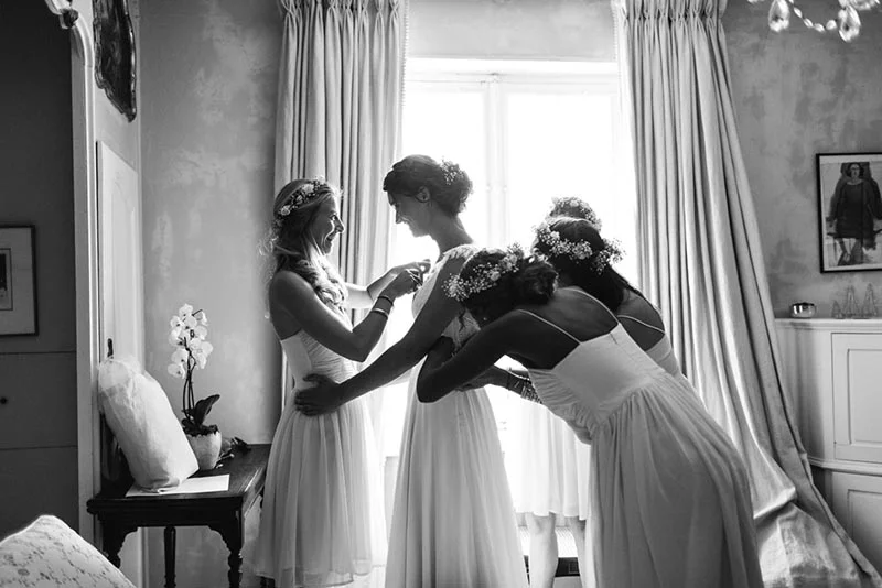 Photo noir et blanc de la mariée entourée de ses demoiselles d'honneur en robes blanches dans une chambre lumineuse.