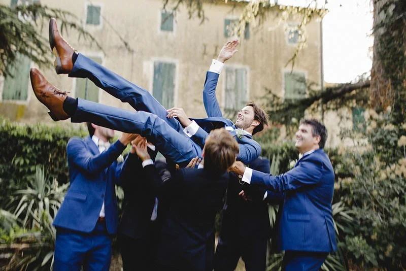 Un groupe d'hommes en costumes bleus soulève joyeusement le marié dans un jardin devant un bâtiment ancien.