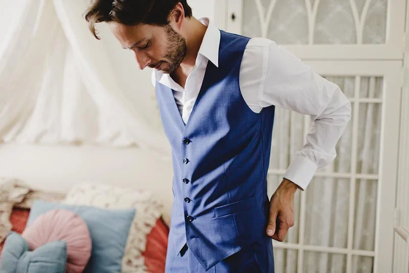 Homme élégant en gilet bleu et chemise blanche s'apprêtant pour son mariage dans une chambre lumineuse.