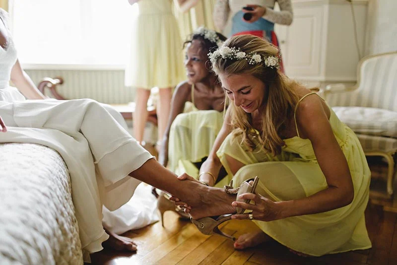 Une demoiselle d'honneur en robe jaune aide la mariée à mettre ses chaussures dans une ambiance douce et lumineuse.