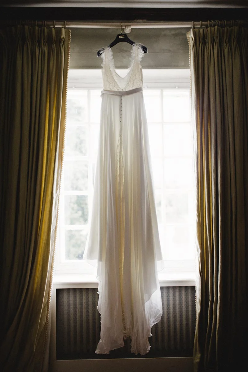 Robe de mariée blanche et fluide suspendue devant une fenêtre lumineuse, encadrée par des rideaux dorés.