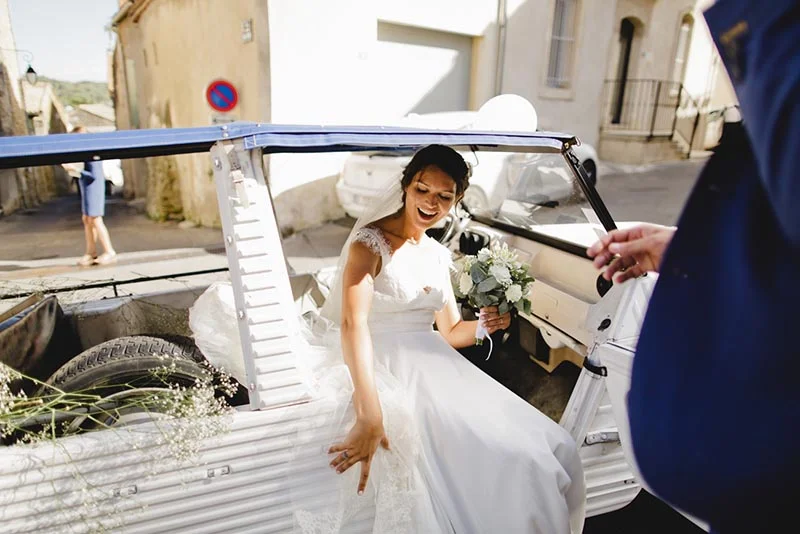 Une mariée souriante en robe blanche descend d'une voiture ancienne décorée dans une rue ensoleillée.