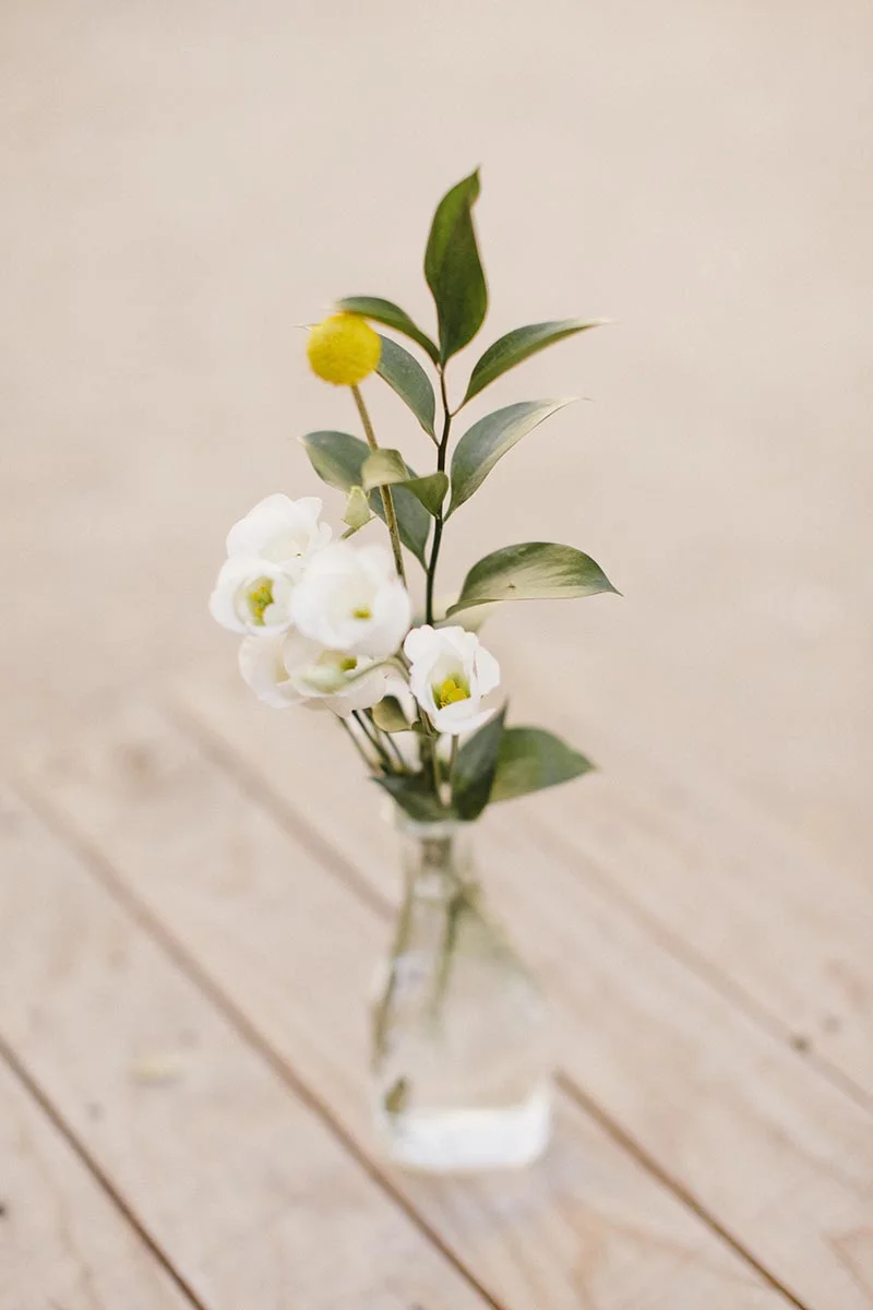 Petit bouquet de fleurs blanches et d'une Craspedia jaune dans un vase en verre sur une table en bois clair.