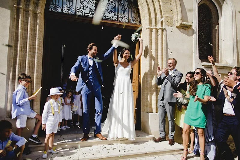 Jeunes mariés joyeux sortant d'une église sous une pluie de confettis, entourés de leurs invités et enfants.