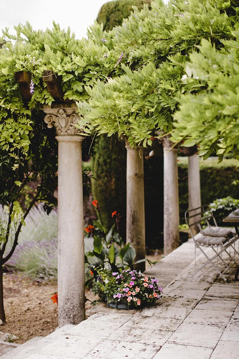 Allée romantique en pierre bordée de colonnes classiques et d'une pergola fleurie sous un feuillage verdoyant.