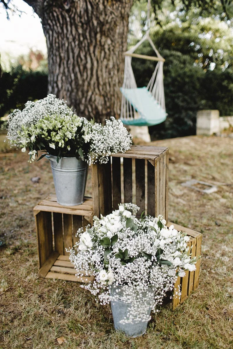 Décoration champêtre avec bouquets de gypsophile blancs dans des seaux zinc et caisses en bois sous un grand arbre.