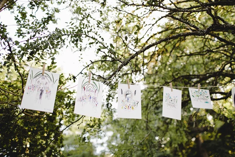 Illustrations de mariage suspendues à une cordelette entre des arbres verdoyants pour une décoration champêtre et poétique.