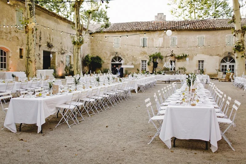 Longues tables dressées en blanc pour un dîner de mariage en plein air dans la cour d'un mas provençal.