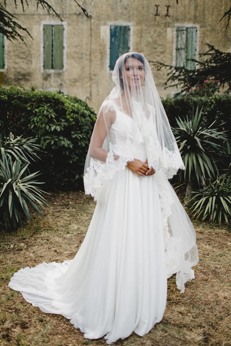 Mariée en robe blanche fluide et voile en dentelle, posant devant un vieux bâtiment en pierre et une végétation verdoyante.