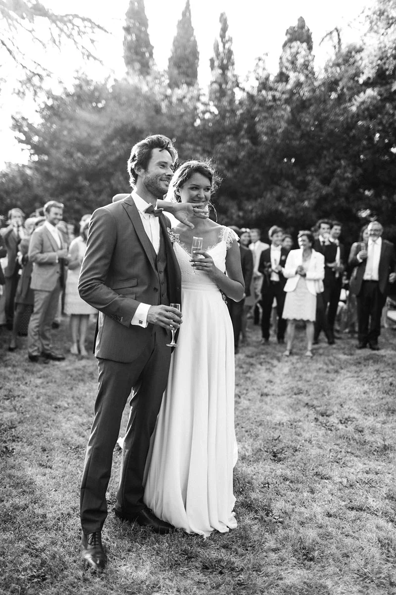 Couple de mariés souriants en noir et blanc lors d'une réception en plein air entourés d'invités.