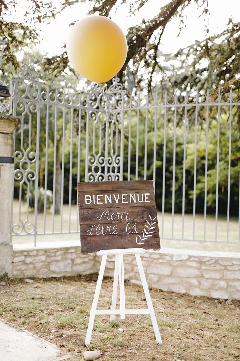 Panneau de bienvenue en bois sur chevalet blanc avec un ballon jaune, devant une grille ancienne et un jardin.
