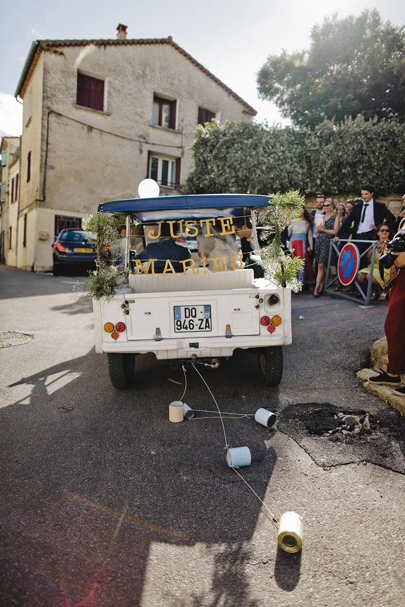 Une voiturette blanche décorée « Juste Mariés » avec des boîtes de conserve, entourée d'invités dans un village.