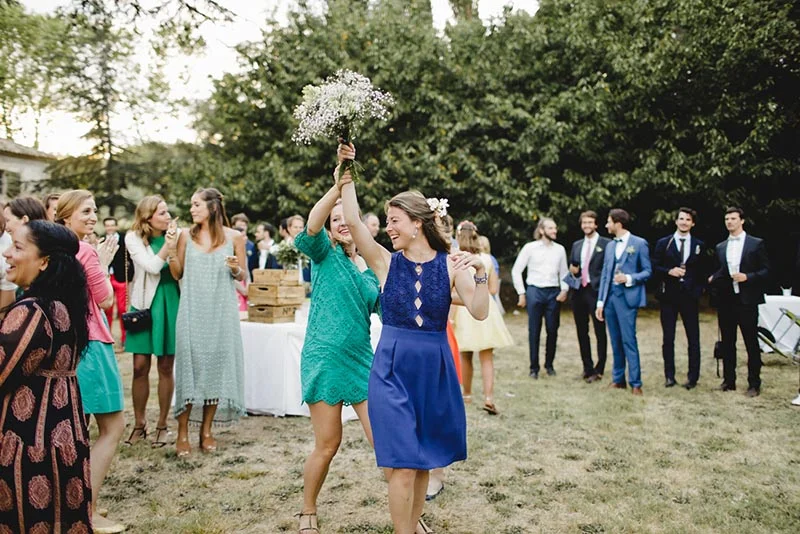 Deux femmes rient et célèbrent en levant un bouquet de fleurs blanches lors d'un mariage en plein air.