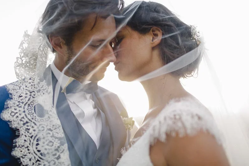 Couple de mariés s'embrassant tendrement sous un voile blanc vaporeux dans une ambiance lumineuse et romantique.