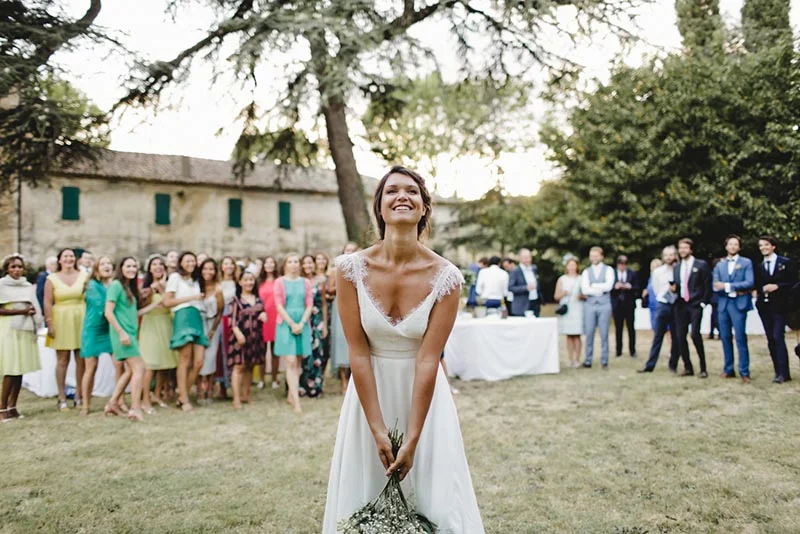 Une mariée souriante en robe blanche avec son bouquet, entourée de ses invités dans un jardin ensoleillé.