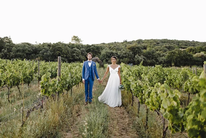 Jeunes mariés main dans la main dans un vignoble, elle en robe blanche et lui en costume bleu, ambiance champêtre.