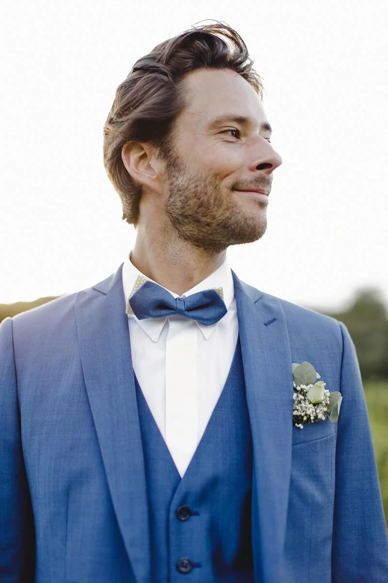 Homme souriant en costume trois-pièces bleu avec nœud papillon et boutonnière fleurie pour son mariage.