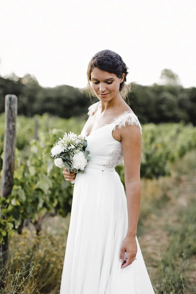 Mariée en robe blanche bohème tenant un bouquet, posant avec douceur dans un vignoble verdoyant.