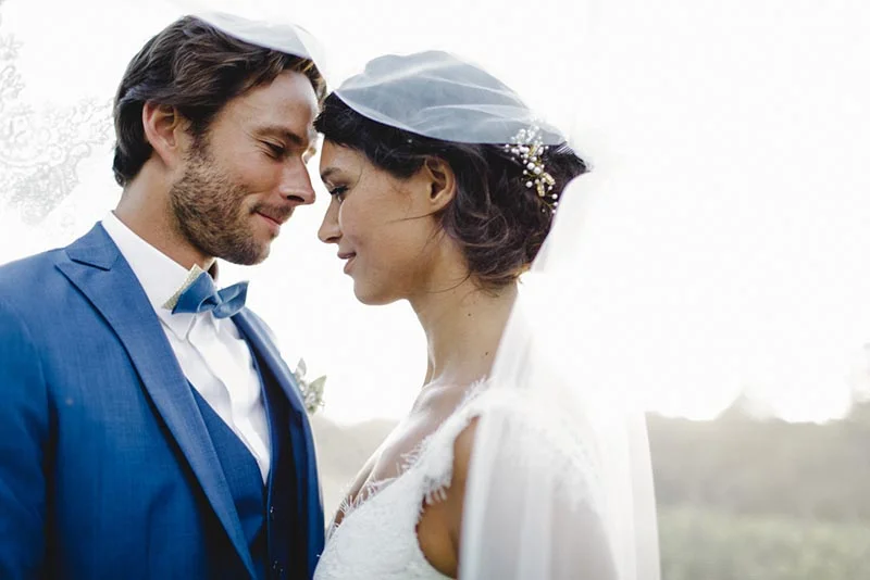 Jeunes mariés complices, lui en costume bleu et elle en robe blanche avec voile, se regardant tendrement en extérieur.