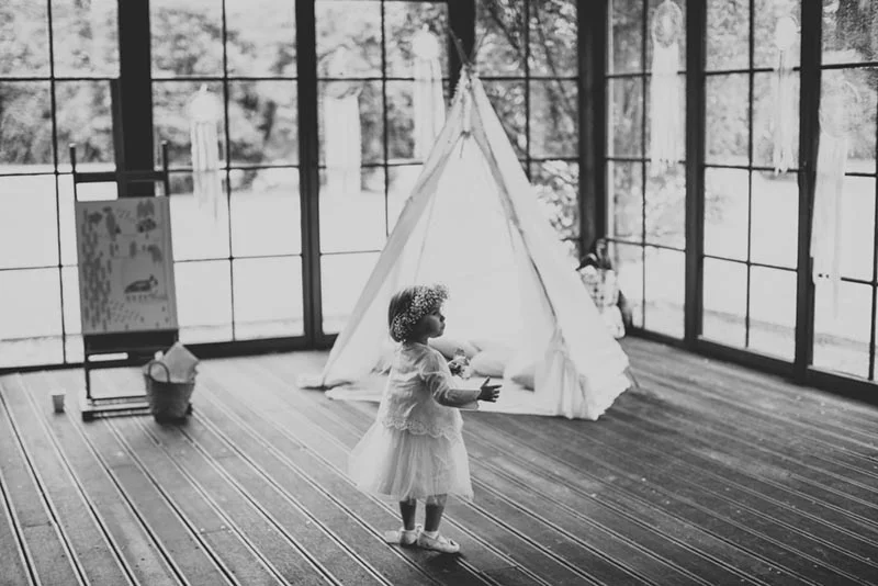 Petite fille en robe blanche et couronne de fleurs devant un tipi, photo en noir et blanc à l'ambiance poétique.