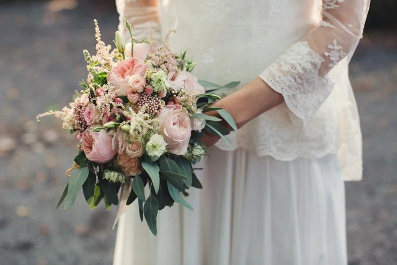 Mariée en robe de dentelle blanche tenant un bouquet romantique de roses roses et de feuillage vert.
