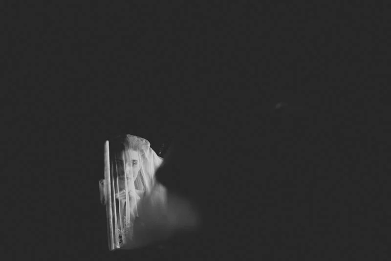 Portrait artistique en noir et blanc d'une mariée voilée, émergeant d'un fond sombre et minimaliste.