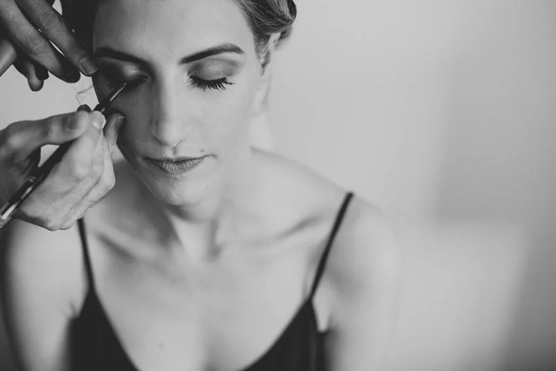 Portrait en noir et blanc d'une mariée se faisant maquiller les yeux avec précision.