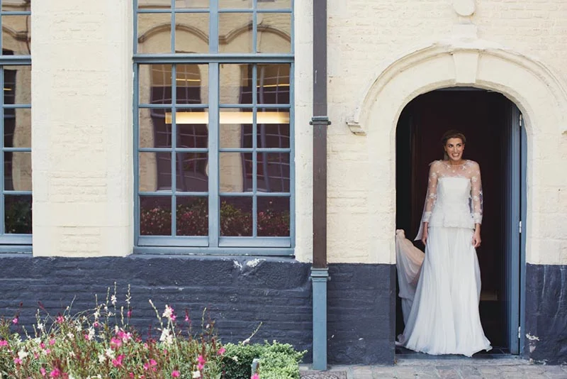 Une mariée en robe blanche et dentelle sortant d'une porte voûtée d'un bâtiment ancien aux murs crème et gris.