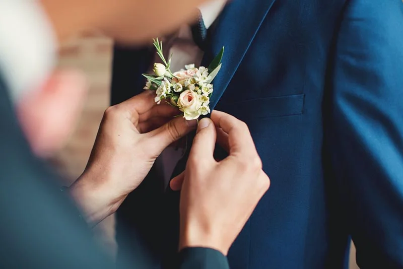 Gros plan de mains ajustant une boutonnière de fleurs blanches sur un costume bleu marine élégant.