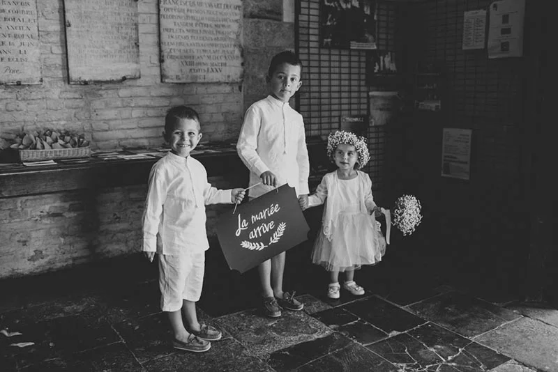Trois enfants d'honneur en tenue blanche posant dans une église ancienne, l'un tenant un panneau La mariée arrive.