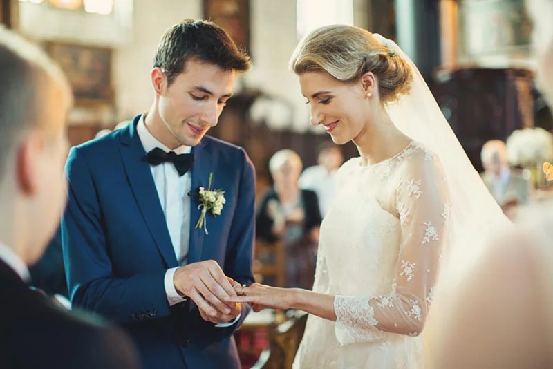 Un marié en costume bleu passe l'alliance au doigt d'une mariée en robe blanche lors d'une cérémonie romantique.