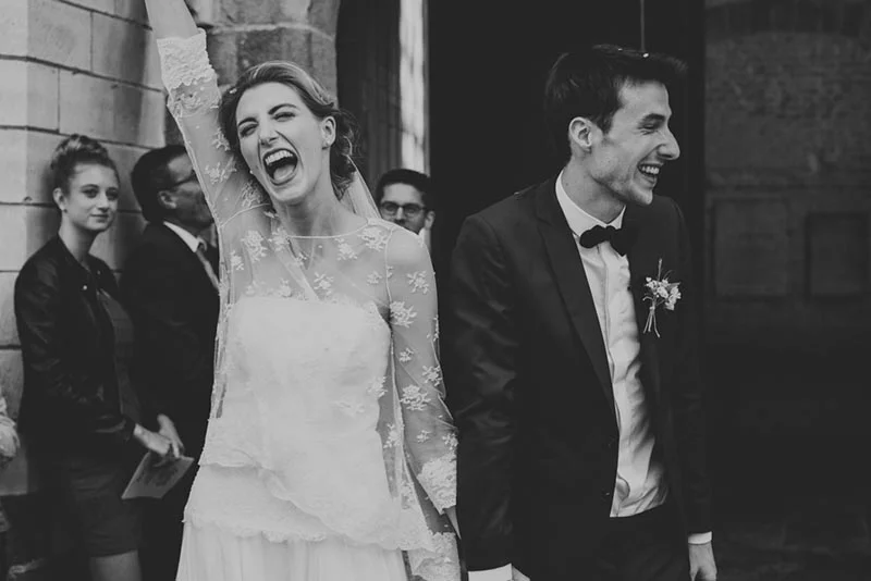 Jeunes mariés riant aux éclats en noir et blanc, la mariée levant le bras avec joie devant des invités.
