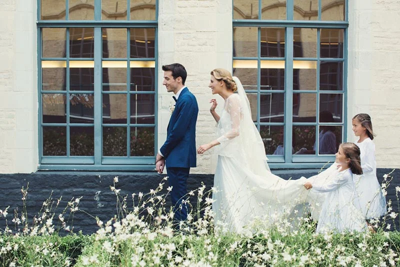 Mariés et demoiselles d'honneur marchant devant une façade blanche et des fenêtres bleues, entourés de fleurs blanches.