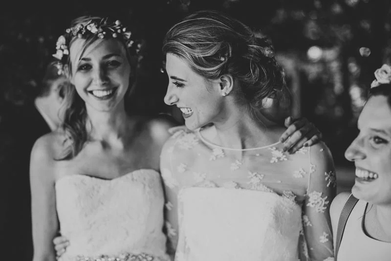 Trois femmes souriantes en robes de mariée blanches, dont l'une porte une couronne de fleurs, en noir et blanc.
