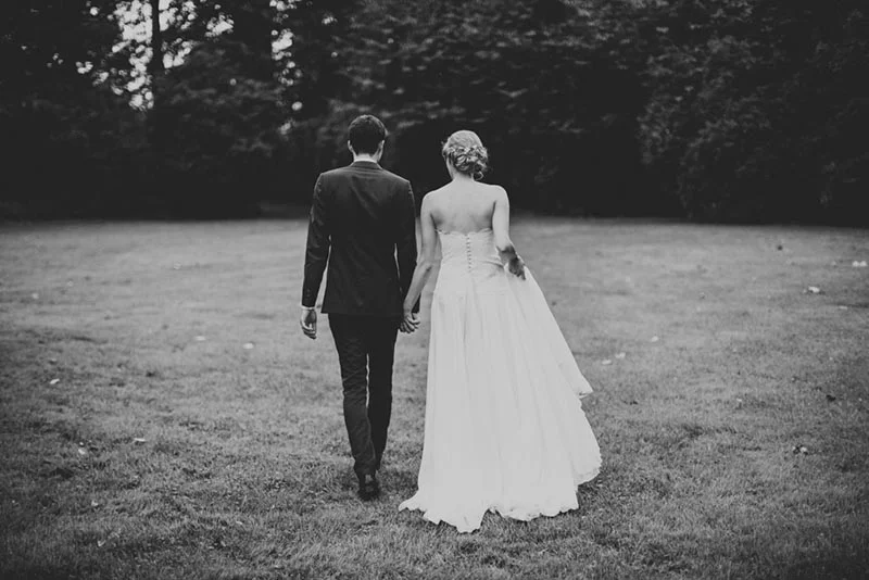 Couple de mariés de dos, main dans la main, marchant dans un champ en noir et blanc.