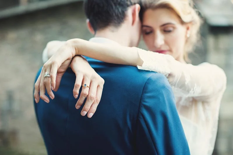 Une mariée en robe blanche enlace un marié en costume bleu, mettant en avant leurs alliances.