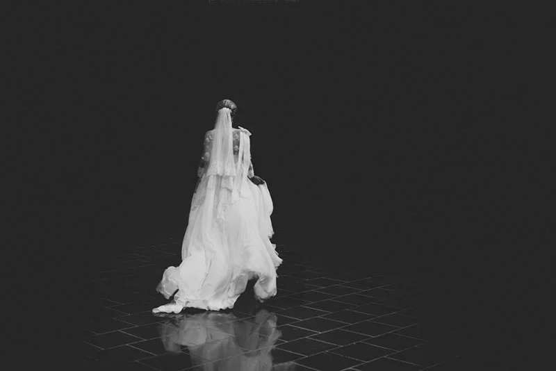 Mariée de dos en robe blanche et voile, seule dans un espace sombre et minimaliste, photographie en noir et blanc.