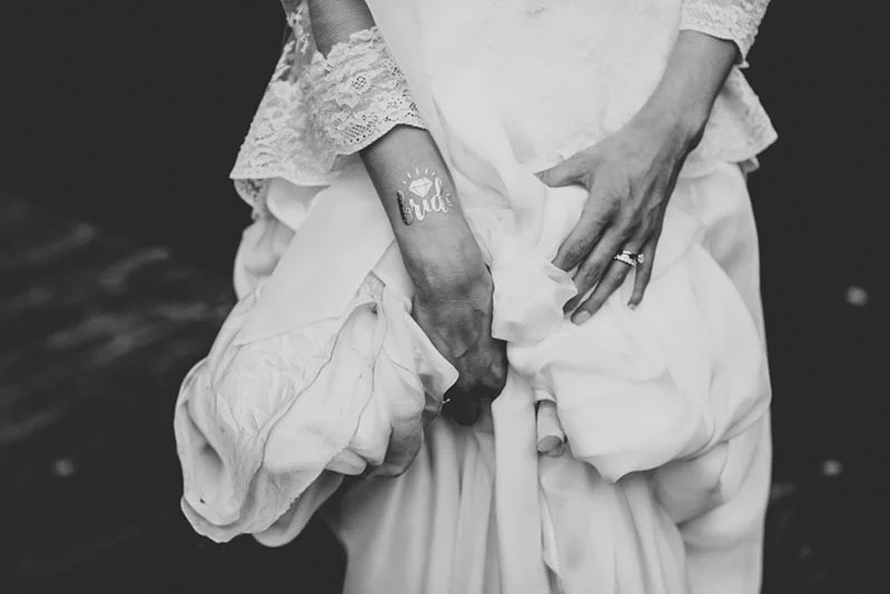 Gros plan en noir et blanc d'une mariée tenant sa robe, avec un tatouage temporaire bride sur le poignet.