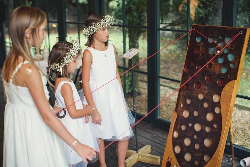 Trois petites filles en robes blanches et couronnes de fleurs s'amusent avec un jeu en bois lors d'un mariage.