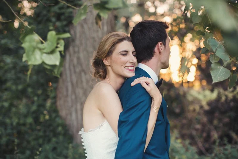 Jeune mariée souriante en robe blanche enlace son époux en costume bleu dans un cadre naturel et lumineux.