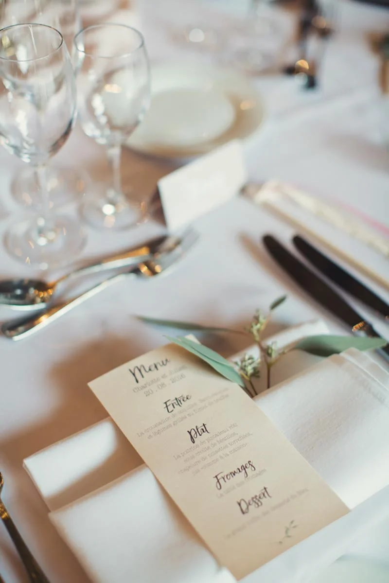 Détail d'une table de mariage élégante avec menu beige, serviettes blanches et verres à pied dans des tons clairs.