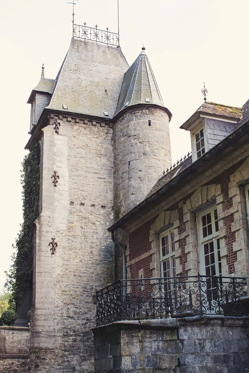 Tour médiévale en pierre claire et bâtiment ancien avec balcon en fer forgé sous un ciel pâle.