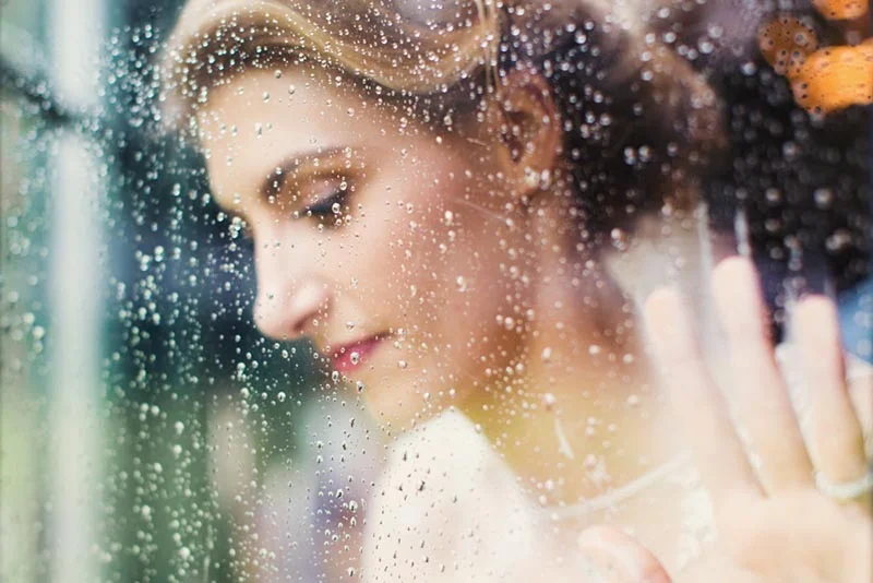 Portrait mélancolique d'une mariée vue à travers une vitre couverte de gouttes de pluie.